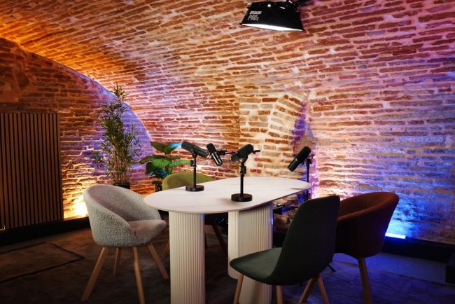 Table ovale, quatre chaises et trois micros dans le décor briques rouges du studio La Voûte